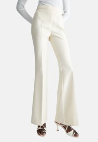 Pantaloni a campana color crema con una finitura liscia, fronte piatto e cintura; abbinati a sandali con tacco aperti bianchi.