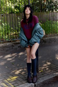 Model purtând o bluză cu mâneci lungi, cu dungi roșii și bleumarin, geacă puf verde, pantaloni scurți din denim, șosete gri până la genunchi și saboți maro.