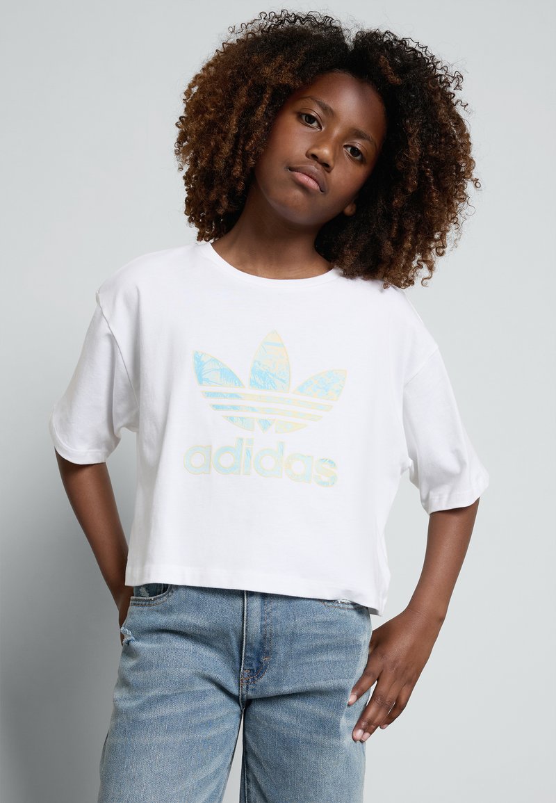 T-shirt cropped branca feito de algodão com um grande logotipo iridescente da Adidas ao centro, combinado com jeans de denim azul claro.