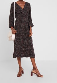 Lang zwart dress met een V-hals, kleine kleurrijke spikkels en uitlopende mouwen. Gecombineerd met bruine sleehak sandalen en een lichte schoudertas.