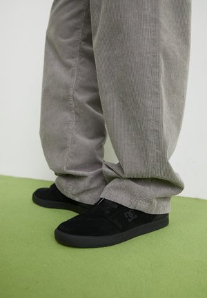 Czarne zamszowe buty do skateboardingu z teksturowanym wykończeniem i logo, w zestawieniu z szarymi spodniami z korduroju o prążkowanej fakturze. Zielona powierzchnia podłogi.