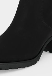 Gros plan d’une botte noire à la cheville montrant l’orteil arrondi, la semelle épaisse avec des rainures profondes et une partie de la tige cousue.