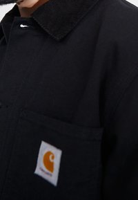 Primo piano di una persona che indossa una giacca nera Carhartt con un colletto visibile e una tasca frontale che mostra l'etichetta del logo bianco e arancione del marchio.