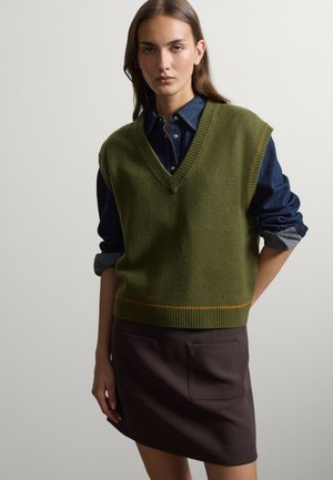 Mujer que lleva un chaleco de punto sin mangas verde sobre una camisa azul oscuro abotonada con las mangas remangadas y una falda marrón oscuro con un bolsillo delantero.