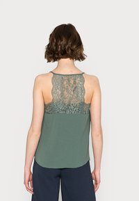 Débardeur vert avec un panneau arrière en dentelle, des bretelles fines et un ourlet arrondi. La texture est douce, combinant des éléments décontractés et élégants.