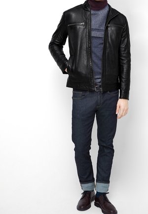 Mand iført sort læderjakke, blå sweater, mørke opsmøgede jeans og brune sko, stående med den ene hånd i lommen.