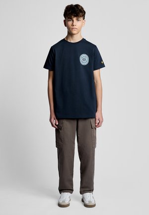 Adolescent debout, portant un t-shirt graphique bleu marine, un pantalon cargo marron et des baskets blanches, sur un fond gris clair uni.