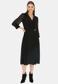 Czarna sukienka z dopasowanym wcięciem w talii, przezroczystymi długimi rękawami i paskiem z okrągłą klamrą. Rozszerzona spódnica i czarne botki do kostek.