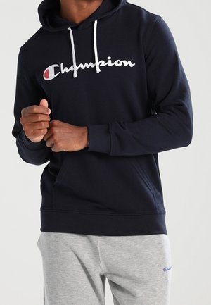 Man die een marineblauwe Champion-trui met wit logo en grijze Champion-sweatpants draagt, en staat met handen dicht bij de borst.