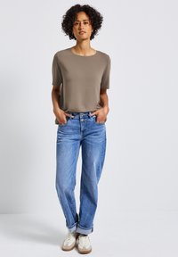 Lehký, taupe tričko s krátkým rukávem v kombinaci s světle modrými, volně střiženými džínami a krémovými teniskami, ukazující neformální styl.