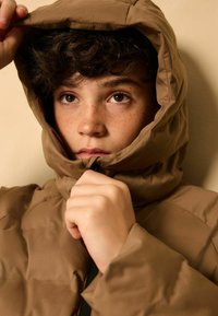 Veste isolante marron avec capuche. Le matériau est lisse et légèrement brillant. Elle possède une fermeture éclair à l'avant et est portée partiellement ouverte, révélant les mains.