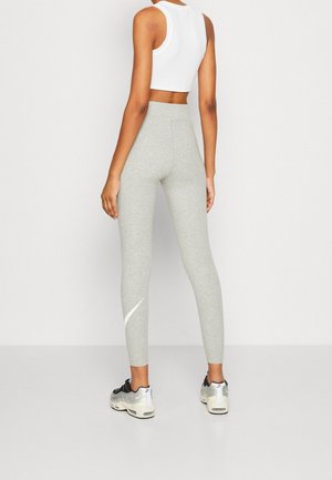 Modelo femenina con leggings gris claro con una franja diagonal blanca cerca de la pantorrilla, top corto blanco y zapatillas blancas y negras, de espaldas.