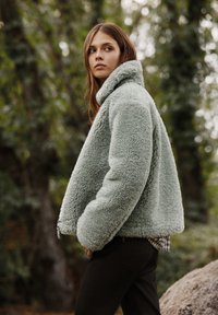 Vihreä, pörröinen shearling-takki korkealla kauluksella, ylisuurena ja sivutaskuilla; yhdistetty tummaan housupariin ja ruudulliseen paitaan alla.