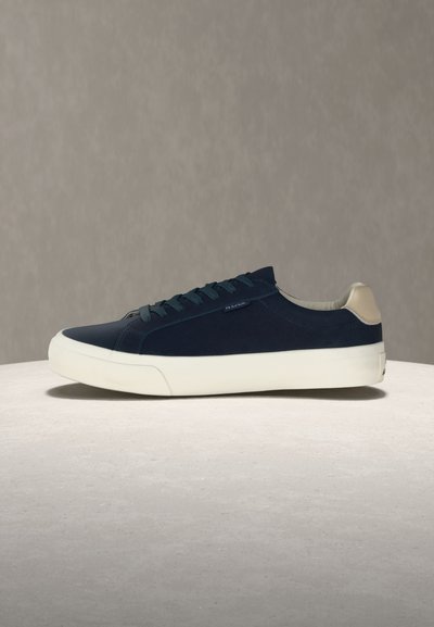 Ναυτικό μπλε χαμηλό αθλητικό παπούτσι με λευκή σόλα και μπεζ λεπτομέρεια στη φτέρνα, πλευρική όψη, σε ανοιχτή επιφάνεια με απλό γκρι φόντο.