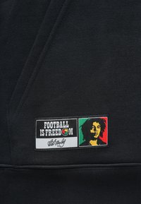 Μαύρο φούτερ με υφασμάτινο μπάλωμα που φέρει το κείμενο "Το ποδόσφαιρο είναι ελευθερία" και ένα πολύχρωμο πορτρέτο ενός άνδρα, πιθανώς του Μπομπ Μάρλεϊ.