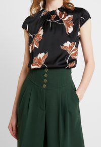 Sort bluse med blomstermønster i brune og creme nuancer, med en rynket halsudskæring og korte ærmer. Sammen med højtaljede grønne bukser med knapper.