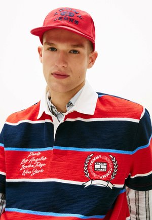 Joven con gorra roja de Tommy Jeans y camiseta de rugby Tommy Jeans a rayas en rojo, azul marino, blanco y azul, con nombres de ciudades bordados.