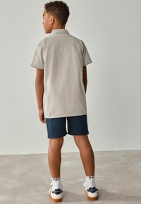 Kurzärmliges hellgraues Hemd mit gestreifter Textur, kombiniert mit dunkelblauen Shorts. Sneaker mit marinefarbenen Akzenten und weißen Socken mit "BAKER"-Logo.