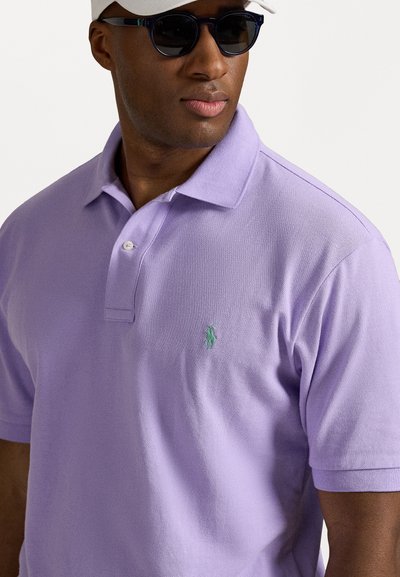 Gai violet polo krekls, izgatavots no teksturēta auduma, ar divu pogu aizdari un mazu zaļu izšūtu logotipu uz kreisā krūša.