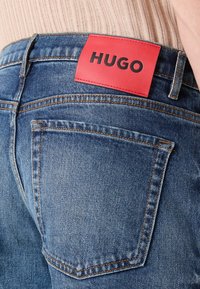 Un bolsillo de pantalones vaqueros azul de mezclilla con costuras naranjas y una etiqueta de cuero rojo con la inscripción "HUGO" en la cinturilla, combinado con una blusa de punto beige.