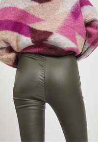 Leggings ecologici in pelle sintetica verde con una texture liscia e dettagli di cucitura distintivi, abbinati a un maglione lavorato a maglia con un motivo multicolore.