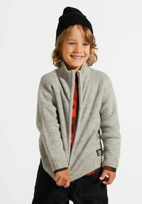 Szary prążkowany sweter z wysokim kołnierzem, zestawiony z czerwoną i czarną koszulą w kratę. Czarna dziana czapka oraz czarne sztruksowe spodnie.