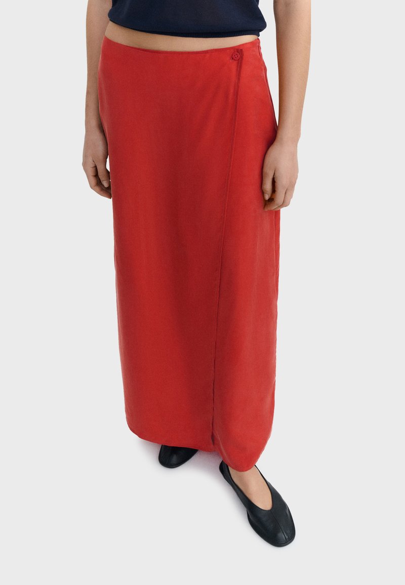 Donna che indossa una lunga gonna avvolgente di un rosso brillante con chiusura a bottoni, abbinata a scarpe nere senza lacci e a un top corto scuro.