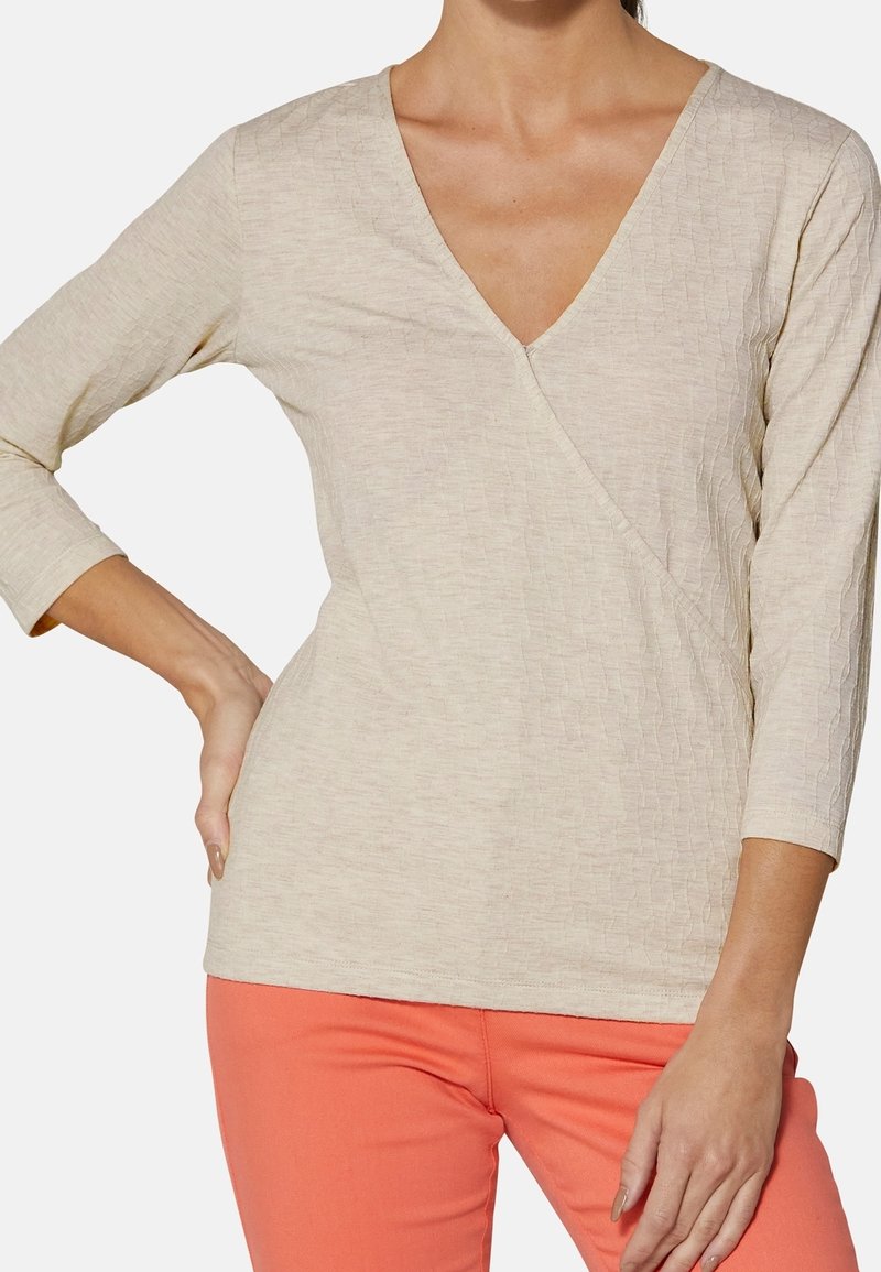 Beżowy, teksturowany top z długim rękawem i dekoltem w kształcie litery V. Posiada styl kopertowy oraz subtelny wzór, zestawiony z koralowymi spodniami.