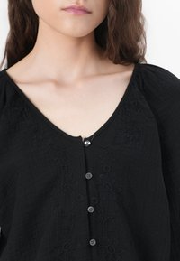 Black cotton blouse with a V-neck, featuring floral embroidery and round buttons down the front. Textured fabric with puffed sleeves.