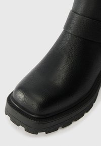 Crne čizme od umjetne kože s zaobljenim vrhom, teksturiranom površinom i debelim potplatom s izraženim uzorcima za hodanje. Traka za naglašavanje u blizini članka.