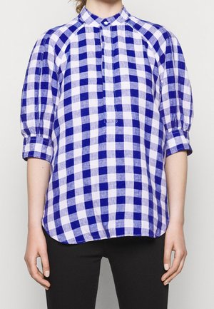 Mujer con blusa de cuadros azul y blanca con mangas abullonadas hasta el codo y cuello alto, combinada con pantalones negros.