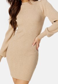 Robe côtelée moulante de couleur beige. Elle comporte des manches longues avec des épaules bouffantes et un col montant. Texture douce et longueur jusqu'aux genoux.