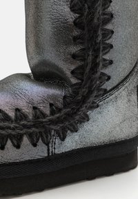 Botín de tobillo plateado metálico con superficie texturizada, costuras decorativas negras, acentos trenzados y suela de goma plana.