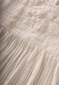 Tissu beige clair avec une surface texturée et froissée. Présente une broderie florale tonale et des accents de dentelle complexes sur l'ensemble du design.