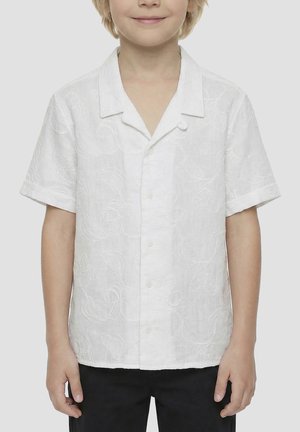 Bambino che indossa una camicia bianca a maniche corte con ricamo floreale sottile, in piedi con le braccia rilassate lungo i fianchi.