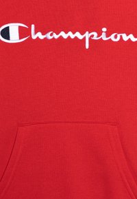 Tessuto rosso con logo "Champion" ricamato in bianco e un emblema in bianco e nero sopra, al di sopra della cucitura della tasca a canguro.