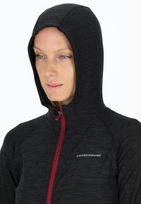 Black hooded jacket with red zipper and logo. Features textured fabric with a fitted design, showcasing a close-up view of the neckline and hood.
