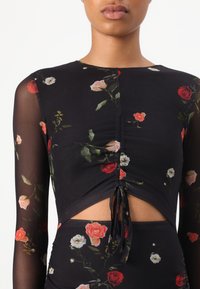 Vrouw draagt een zwarte bloemenmesh top met lange mouwen en een gerimpeld voorstuk met strik, over bijpassende hoog getailleerde bloemenbroeken.
