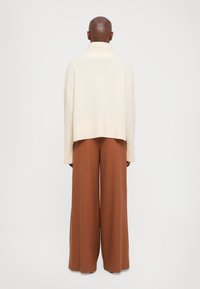 Łysy mężczyzna w kremowym swetrze z golfem i szerokich brązowych spodniach, stojący tyłem na jednolitym białym tle.