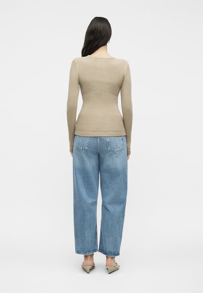 Γυναίκα όρθια με την πλάτη, φοράει μπεζ ριμπ πλεκτό μακρυμάνικο τοπ, φαρδιά μπλε τζιν και μπεζ σανδάλια με τακούνι σε μονόχρωμο φόντο.