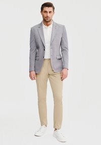 Jasnoszary blazer z klapami, dwoma guzikami i bocznymi kieszeniami, w połączeniu z białą koszulką polo i beżowymi spodniami, dopełniony białymi sneakersami.