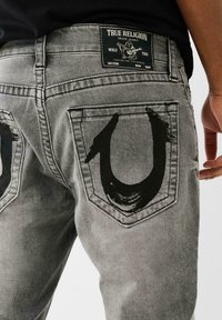 Jeans din denim gri, cu un logo stilizat negru pe buzunarele din spate, având o etichetă neagră pe talie pe care scrie "True Religion".