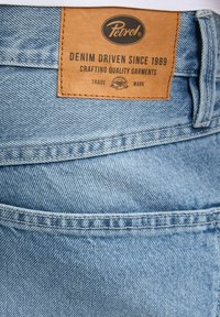 Jasnoniebieska tkanina dżinsowa z naszywką ze skóry oznaczoną napisem "Petrol", zawierająca tekst: "DENIM DRIVEN SINCE 1989" oraz widoczne detale szwów.