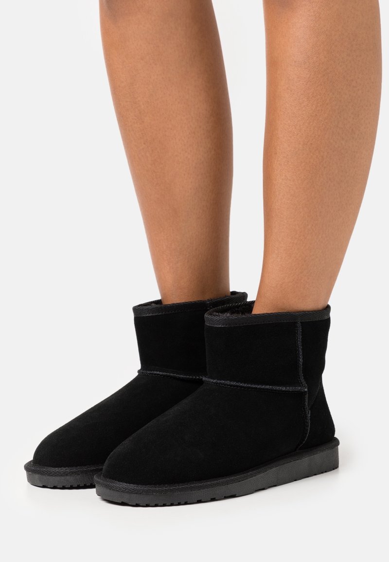 Pieds chaussés de bottes en suède noir mi-mollet avec des semelles épaisses, présentées sur un fond clair uni.