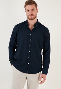Camisa de botón de manga larga texturizada en azul marino con cuello puntiagudo y botones blancos. Combinada con pantalones de color claro, mostrando un ajuste relajado.