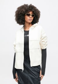 Ελεφαντόδοντο μπουφάν με φερμουάρ και κουκούλα, με πλευρικές τσέπες και ριμπ μανσέτες. Φοριέται πάνω από μαύρο εφαρμοστό πουκάμισο, σετ με μαύρο σορτς.