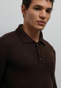 Jonge man met kort donker haar, gekleed in een donkerbruin gebreide trui met een kraag en drie knopen, tegen een effen achtergrond.