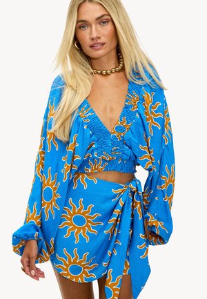 Haut court bleu et jupe drapée ornés de motifs solaires en orange. Le matériau semble léger avec une finition texturée et des manches fluides.