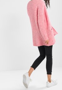 Light pink knitted long cardigan with a loose fit and side pockets, paired with black skinny jeans and white sneakers.