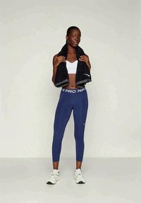 Niebieskie legginsy Nike Pro z białym paskiem w talii, zestawione z białym sportowym biustonoszem. Modelka trzyma czarny ręcznik. Białe buty sportowe.
